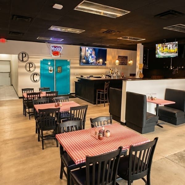 Spacious restaurant interior with red and white checkered tables, black chairs, booths, a bar, and TVs.