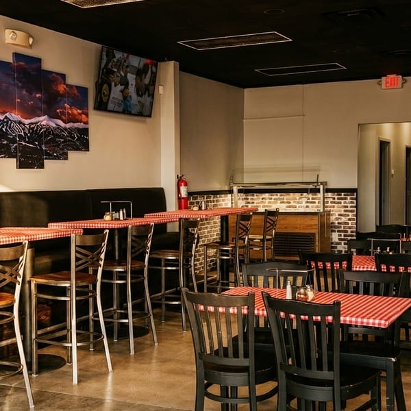 Empty restaurant dining room with red and white checkered tables, black chairs, and mountain artwork.